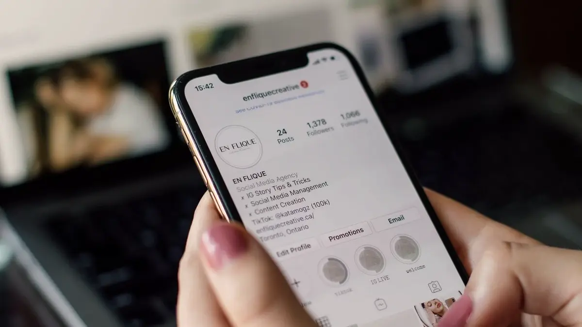 Une main tient un smartphone affichant un profil Instagram, un ordinateur portable flou à l'arrière-plan.