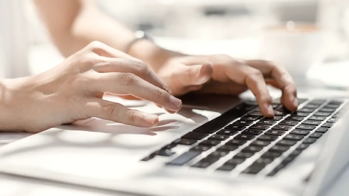 Deux mains tapent sur le clavier d'un ordinateur portable, en contre-jour sur un bureau blanc.