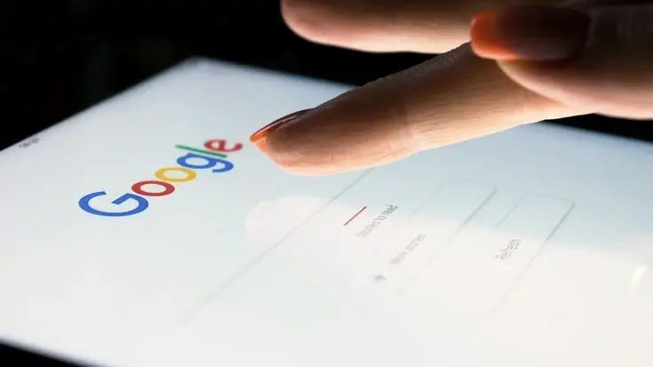 Un doigt s'approche de la page d'accueil de Google affichée sur une tablette, dans une pièce sombre.